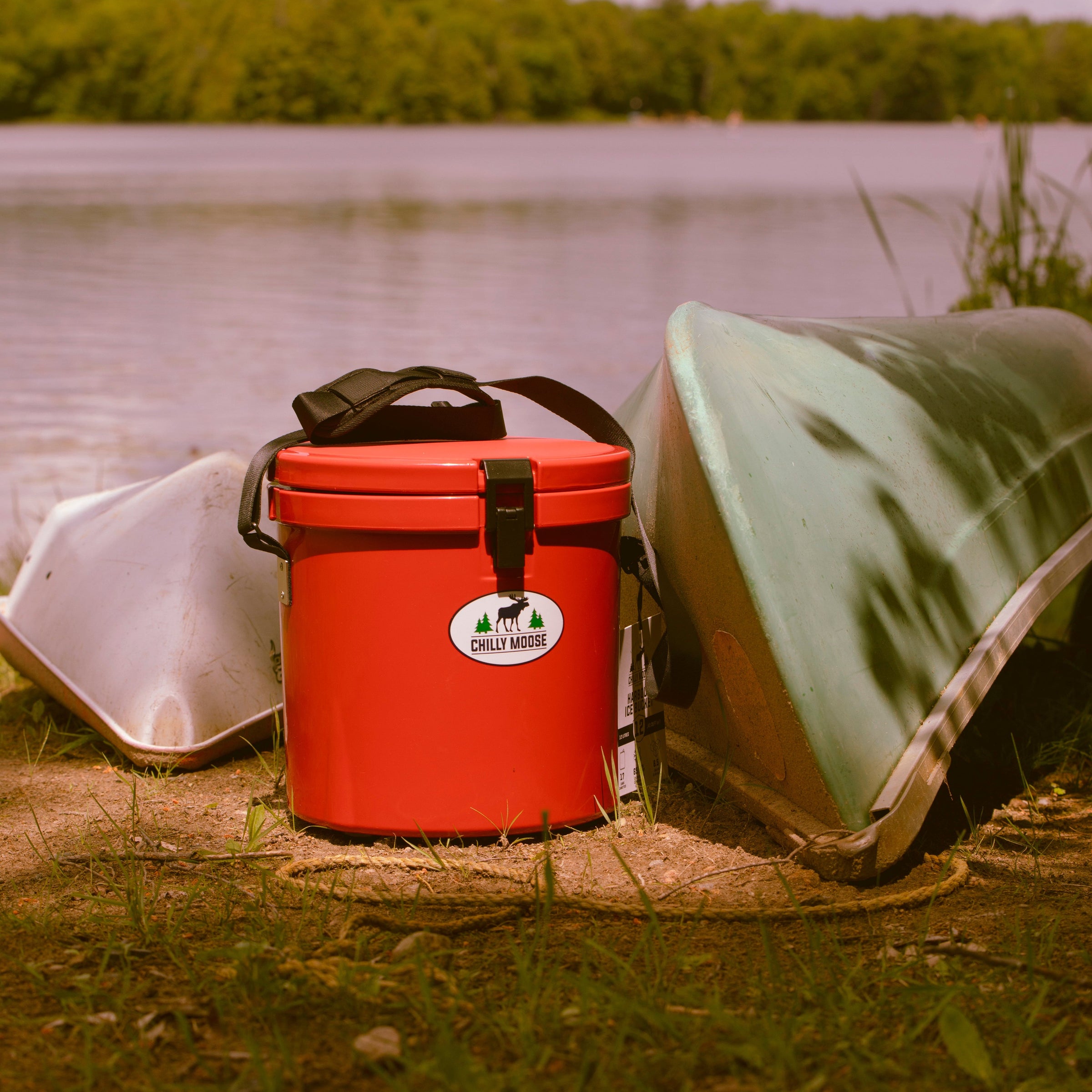 Chilly Moose premium outdoor coolers. Pictured: Chilly Moose 12L Harbour Bucket in Canoe Red and 25L Chilly Ice Box in Canoe Red. Both cooler lids are slightly open. Coolers are positioned on a lakeside dock.