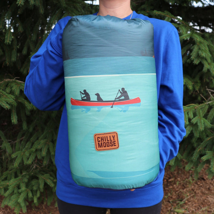 Stillwater Ultralight Puffy Blanket – Chilly Moose – Rolled in stuff sack outdoors, canoe graphic visible #colour_tofino