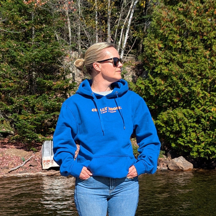 Person wearing a Camp blue hoodie and jeans standing on a wooden dock by a lake with trees in the background.